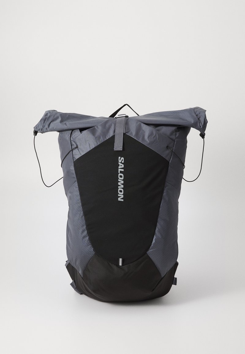 Mochila cinza com design de abertura superior, apresentando um painel preto, superfície texturizada e um cordão ajustável. Logótipo "Salomon" na frente.