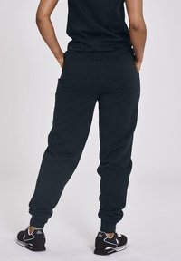 Pantaloni sportivi neri con polsini elasticizzati, tasche laterali e una texture morbida. Indossati con una maglia coordinata e sneaker nere, mostrati da dietro.