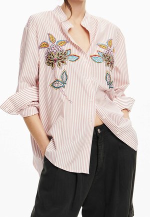 Frau trägt ein locker sitzendes rosa-weiß gestreiftes Hemd mit bunter Blumenstickerei und schwarze Hose, Hände in den Taschen, Ärmel hochgekrempelt.