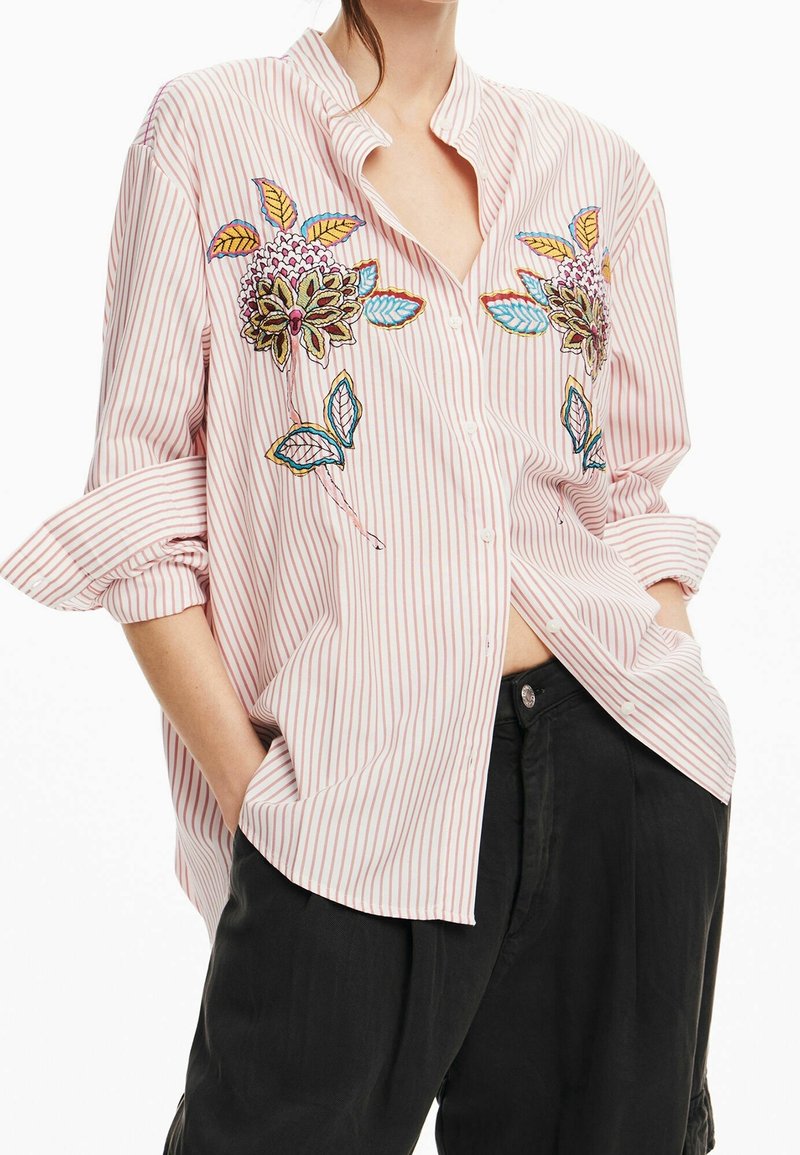 Femme portant une chemise ample à rayures roses et blanches avec broderie florale colorée et pantalon noir, mains dans les poches, manches retroussées.