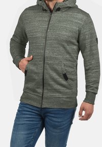 Hombre llevando una sudadera con capucha verde con cremalleras y bolsillos, y vaqueros azules, con la mano en el bolsillo, frente a un fondo liso.