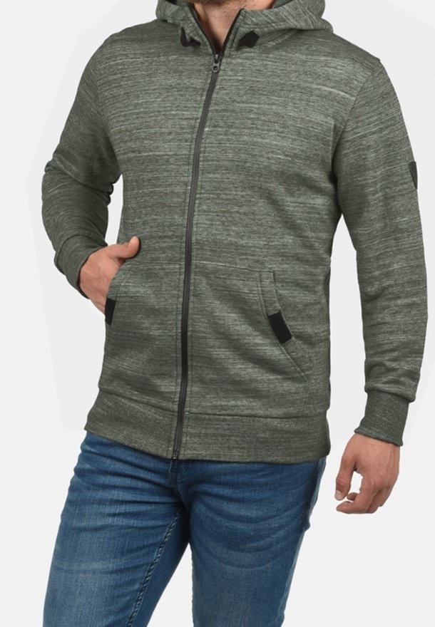 Hombre llevando una sudadera con capucha verde con cremalleras y bolsillos, y vaqueros azules, con la mano en el bolsillo, frente a un fondo liso.