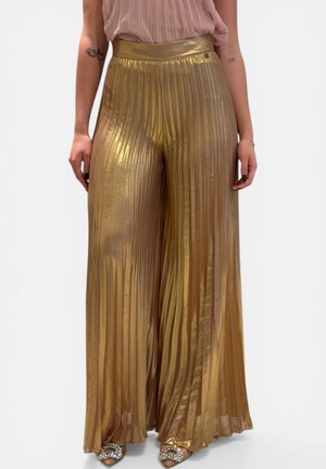 Donna che indossa pantaloni a vita alta in tessuto metallico plissettato color oro a gamba larga, abbinati a una camicetta rosa trasparente e tacchi aperti decorati.