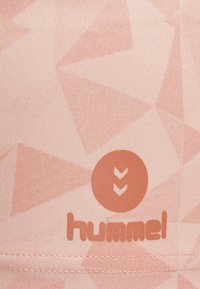 Jasnoróżowa tkanina sportowa z geometrycznym wzorem. Posiada okrągłe logo z pionowymi strzałkami i napisem "hummel" w dopasowanym kolorze.