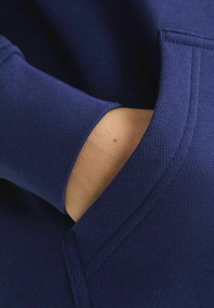 Navyblauer Hoodie aus strukturiertem Stoff mit einer Fronttasche. Nahaufnahme eines Arms, der in der Tasche ruht, zeigt Hautfarbe und ein Muttermal.