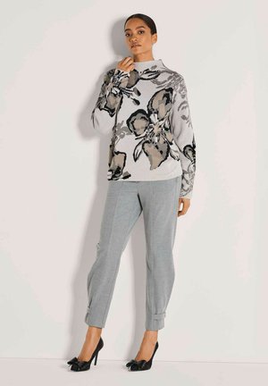 Vrouw die tegen een muur staat, gekleed in een grijze bloemen trui, lichtgrijze broek en zwarte hoge hakken met strikken.