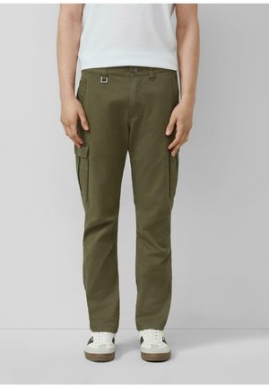 Personne portant un pantalon cargo vert olive avec des poches latérales, des baskets blanches et une chemise blanche, debout devant un fond gris uni.