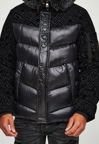 Sort dunjakke med et quiltet design, med en pelsforet krave, præget logo og en blanding af skinnende og tekstureret stof.