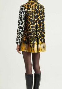 Giacca corta con motivo geometrico di giraffe in nero, bianco e giallo. Tessuto liscio con colletto minimalista e design posteriore plissettato.