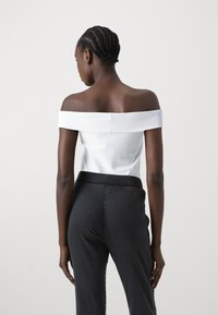 Baltā off-the-shoulder blūze ar vertikālām ribām, kombinēta ar tumši pelēkajām svītrainajām biksēm ar elastīgu jostu un gludu tekstūru.