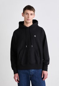 Čierna bavlnená mikina s kapucňou, predným vreckom a logom CK na hrudi. V kombinácii s modrými džínami.