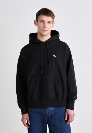 Zwarte katoen hoodie met een voorvak, trekkoordkap, en cK-logo op de borst. Gecombineerd met blauwe denim jeans.