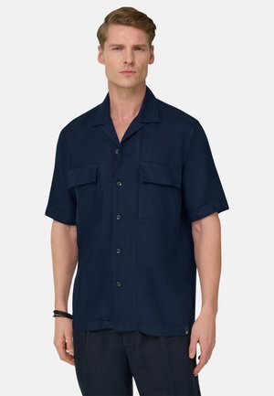 Mies, jolla on päällään tummansininen, lyhythihainen, napitettava paita, jossa on kaksi rintataskua, seisoo vaaleanharmaan taustan edessä.