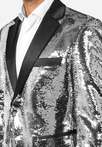 Mężczyzna w błyszczącej srebrnej marynarce z cekinami i czarnymi satynowymi klapami, noszący białą koszulę.