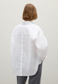 White oversized shirt with a back pleat, classic collar, and buttoned cuffs. Smooth fabric with a matte finish. Gray trousers in view.