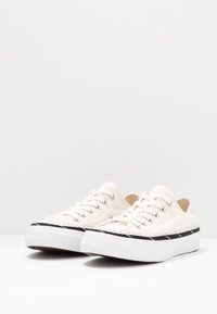 Paire de baskets basses en toile blanche avec lacets blancs et semelles épaisses blanches ornées d'une bande noire près de la base, posées sur une surface blanche.