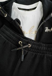 Sudadera negra con cremallera y detalles en dorado, combinada con pantalones negros con cordones que presentan una tela texturizada y detalles del logo en los lazos.