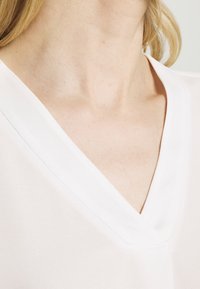 White blouse featuring a V-neckline, soft fabric, and smooth texture. The stitching is subtle, enhancing the garment's clean design.