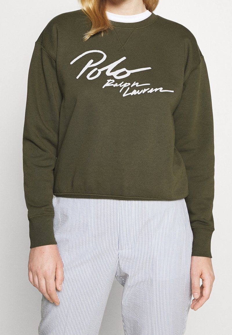 Sweat-shirt vert olive avec inscription blanche "Polo Ralph Lauren", porté avec un pantalon blanc à rayures bleu clair.
