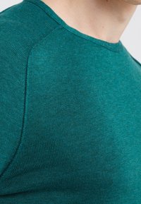 Närbild av en person klädd i en petrolblåstickad tröja med synliga sömmar vid axeln och kragen.