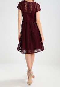 Burgunderfarbenes Spitzenkleid mit tailliertem Oberteil und ausgestellt Rock, ausgestattet mit kurzen Ärmeln und einem Schlüssel-ausschnitt auf der Rückseite. Tragen mit nude-farbenen Absätzen.