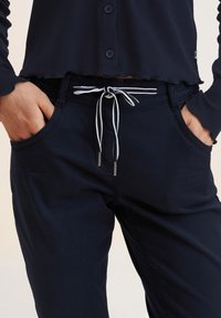 Granatowe spodnie z wiązanym paskiem w pasie, zdobione jasnym obszyciem. Wykonane z miękkiego materiału, posiadają boczne kieszenie i dopasowany krój.