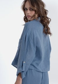 Camicia in lino blu chiaro con una vestibilità rilassata, spalle cadute e maniche arrotolate. Presenta una tasca sul petto e una finitura liscia e textured.
