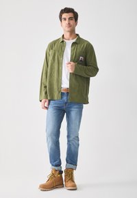 Green corduroy shirt with a front pocket, paired with blue denim jeans and tan lace-up boots. Simple design, relaxed fit, and textured fabric.
