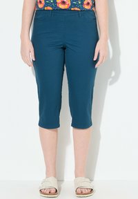 Pantalons capri teal en tissu lisse et extensible avec une taille ajustée, arrivant mi-mollet, associés à des sandales légères.