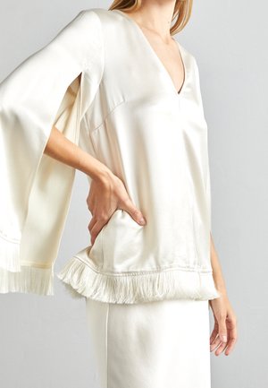 Vrouw draagt een witte satijnen blouse met wijde klokmouwen en franjes aan de mouwen en zoom, gecombineerd met een bijpassende witte rok.