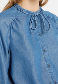 Vaaleansininen farkkupaita, jossa on pitkät hihat, nappisulku ja solmittu rusetti kaulassa, henkilön päällä, joka on kuvattu kaulasta vyötäröön.