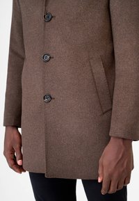 Brun ullblandad kappa med rak skärning, som har knappstängning, två sidofickor och en mjuk textur. Detaljer på knapparna är synliga.