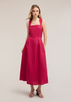 Mujer con vestido midi rosa brillante sin mangas, con escote cuadrado y falda plisada, de pie frente a un fondo neutro.