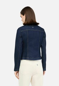 Tmavě modrá denimová bunda s přiléhavým střihem, která má kontrastní stehy a mírně zkrácenou délku. Zadní pohled ukazuje zjednodušený design.