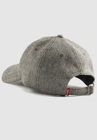 Gorra gris en patrón de espiga con visera curvada, correa ajustable y una etiqueta roja. Tejido texturizado con un prominente patrón y detalles de costura.