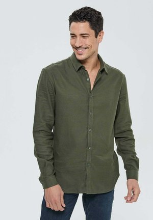 Chemise à manches longues vert olive avec un col boutonné, texture douce et patte de boutonnage sur le devant, portée avec un jean bleu.
