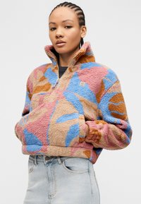 Γυναίκα με πλεξούδες φορώντας ένα πολύχρωμο φλις μπουφάν με αφηρημένο μοτίβο, ψηλό γιακά και τζιν ανοιχτού χρώματος, με τα χέρια στις τσέπες του μπουφάν.