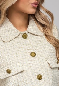 Biała teksturowana kurtka z guzikami w złotym odcieniu, z kołnierzem i klapami na kieszeniach. Tkanina ma tkany wzór dla dodatkowego efektu.