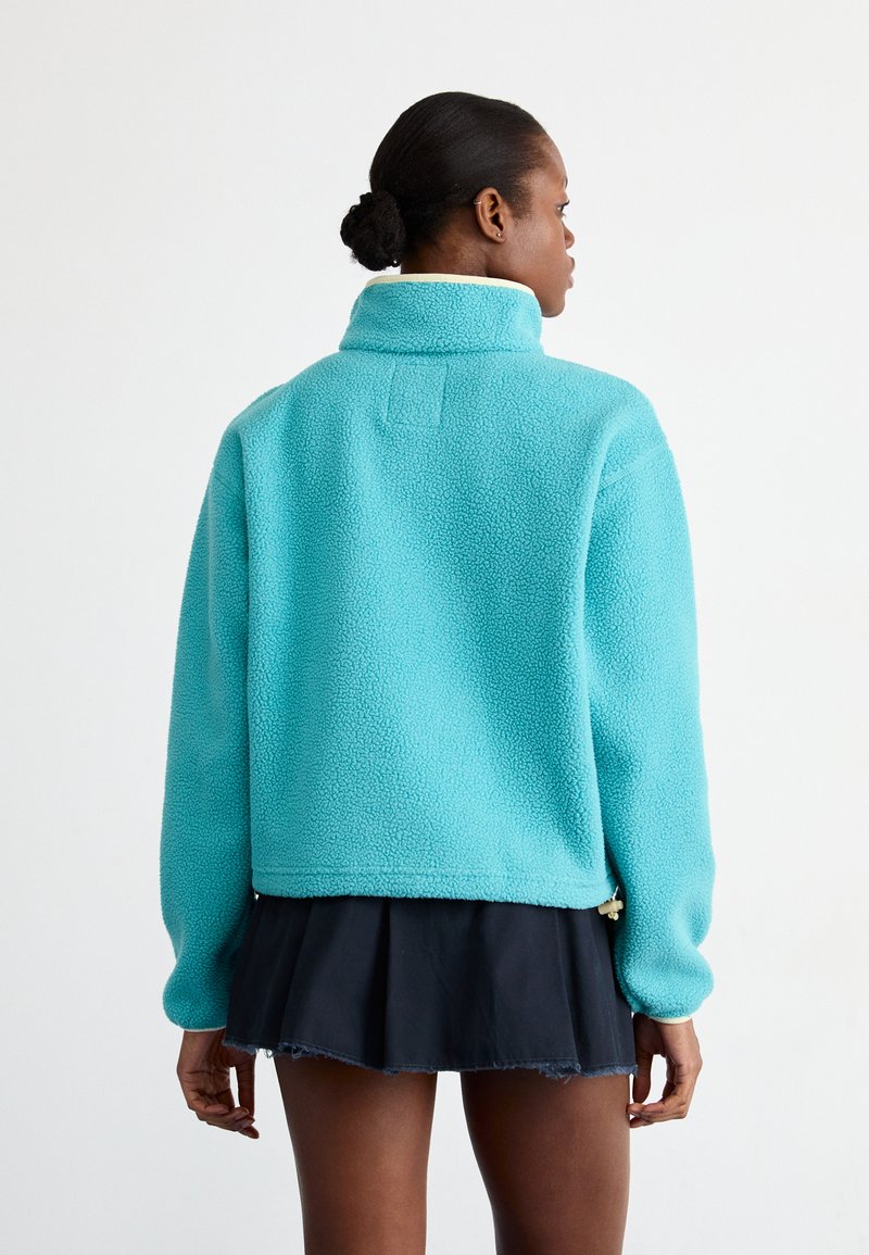 Chaqueta de forro polar turquesa con un alto cuello, hombros caídos y un diseño corto. Presenta un tejido texturizado y detalles de costura visibles.