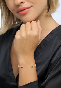 Goldfarbener Armreif mit zwei grünen Edelsteinen, glatte Metalloberfläche, minimalistisches Design, getragen an einer Hand vor einem schwarzen Stoffhintergrund.