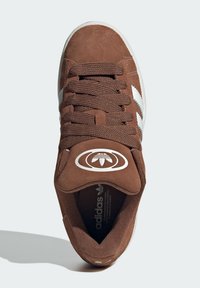 Brown suede sneaker with white stripes, round logo on tongue, lace-up design, textured insole, and rubber outsole. Top view displayed.