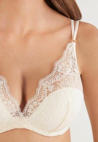 Primer plano de una mujer que lleva un sujetador de encaje color crema con bordados florales y tirantes dobles ajustables.