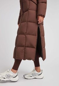 Abrigo inflable marrón con un diseño acolchado, que presenta un zipper lateral y una longitud larga. Usado sobre leggings marrones y zapatillas blancas con acentos plateados.