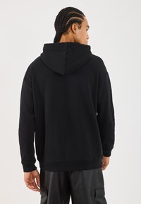 Svart hettegenser laget av mykt materiale, med snor i hetta, ribbede mansjetter og en avslappet passform. Synlig stitchingdetalj langs sømmene.