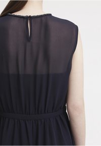 Vestito blu navy con schiena trasparente, caratterizzato da un'apertura a chiave e un collo impreziosito. Il tessuto è morbido e fluido, con una vita aderente.