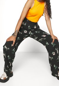 Czarne kwiatowe spodnie z zielonymi i różowymi wzorami, w połączeniu z dopasowanym pomarańczowym topem na ramiączkach. Obuwie ma grubą podeszwę z paskami.
