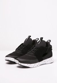 Baskets noires avec une tige en mesh, des sangles élastiques et une semelle texturée avec des taches blanches. Design minimaliste avec un look sportif.