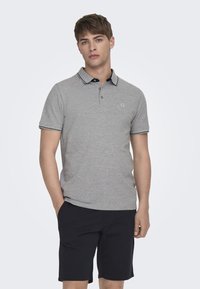 Graues Poloshirt mit schwarzem Kragen und Besatz, kurzen Ärmeln und einem kleinen Logo auf der Brust. Kombiniert mit dunklen Shorts.