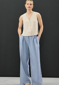 Beige bezručkový top s výstřihem do V a předními knoflíky, spárovaný se světle modrými kalhotami s vysokým pasem a širokými nohavicemi. Texturovaná látka na obou kouscích.