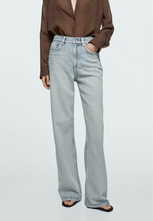 Osoba ubrana w jasnoniebieskie, szerokie dżinsy z wysokim stanem oraz brązową, zapinaną na guziki koszulę włożoną do środka, stojąca z jedną ręką w kieszeni i czarnych sandałach.